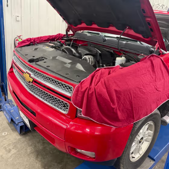 A red Chevrolet SUV with its hood open is on a lift in a garage. Red protective covers are draped over the fenders to prevent scratches during engine maintenance.